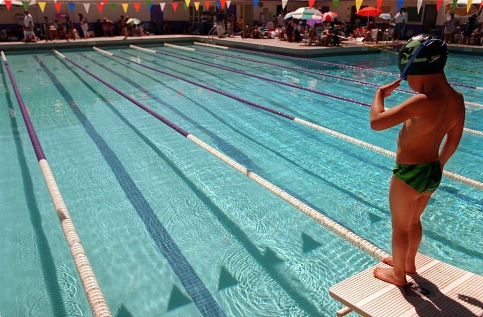 A child in swim cap and swimwear stands on a starting block at the edge of an outdoor pool, preparing for a race. People and umbrellas are seen in the background