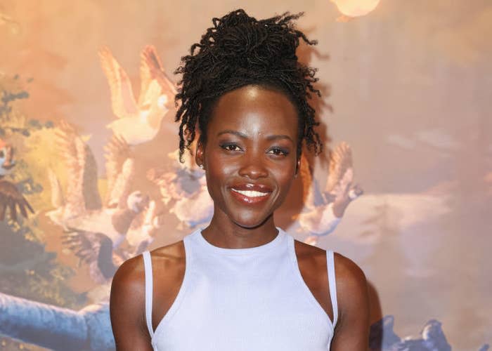 Lupita Nyong'o poses in a sleeveless, fitted dress with a serene bird-themed background