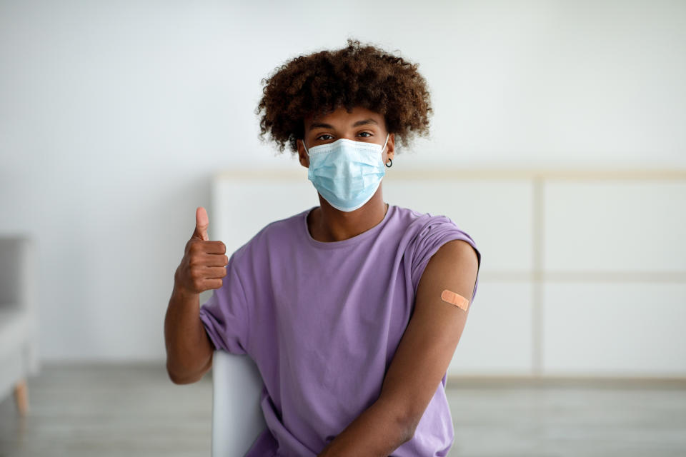 Vaccinated black teen guy in face mask wearing plaster bandage on his arm after covid vaccine injection, showing thumb up at home. African American teenager recommending coronavirus immunization