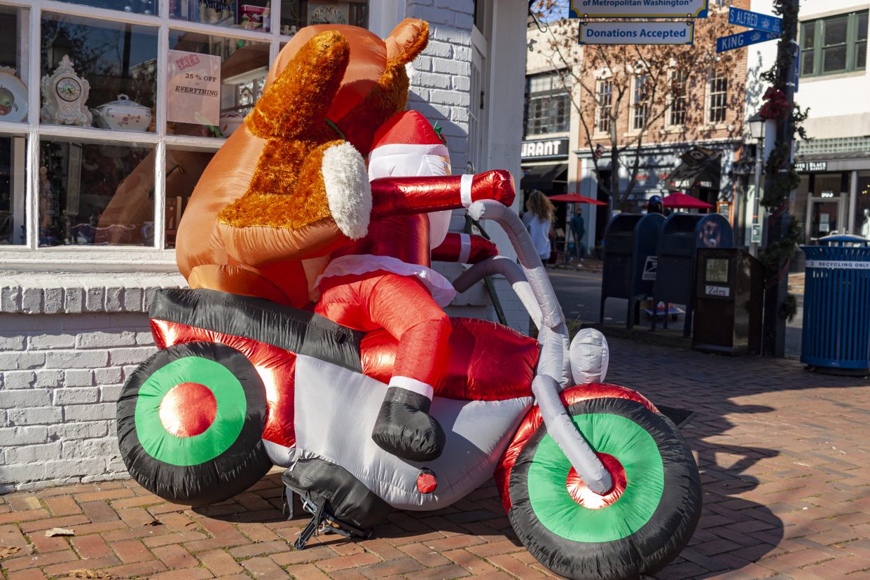 Big inflatable santa claus with bear riding on a motorcycle in downtown, Alexandria, Virginia, during the christmas season, sunlight shining on it, with a shop-lined street in the background