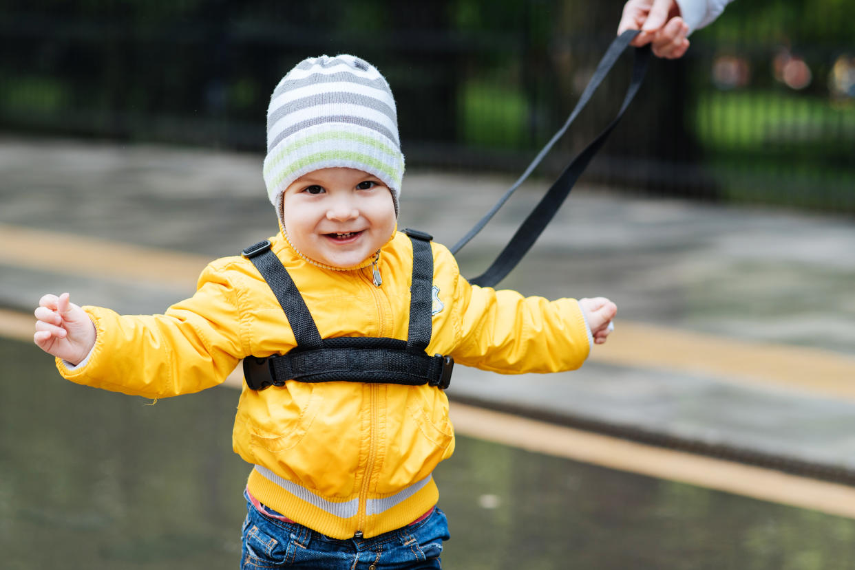 A young child, smiling at the camera, wears a harness and leash. A parent’s hand holds the other end of the leash.