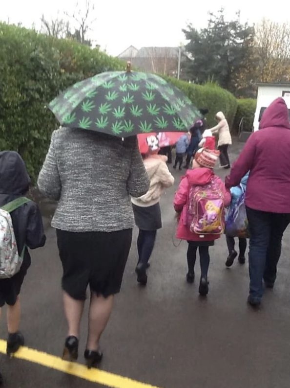 Parents and children walk together, with children carrying school backpacks. An adult holds an umbrella with a leaf pattern, protecting from rain