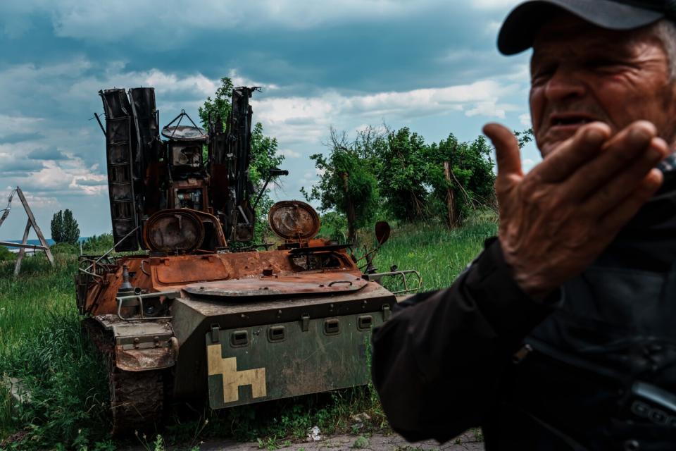 A man gestures near the hulking wreckage of a military vehicle in a grassy area