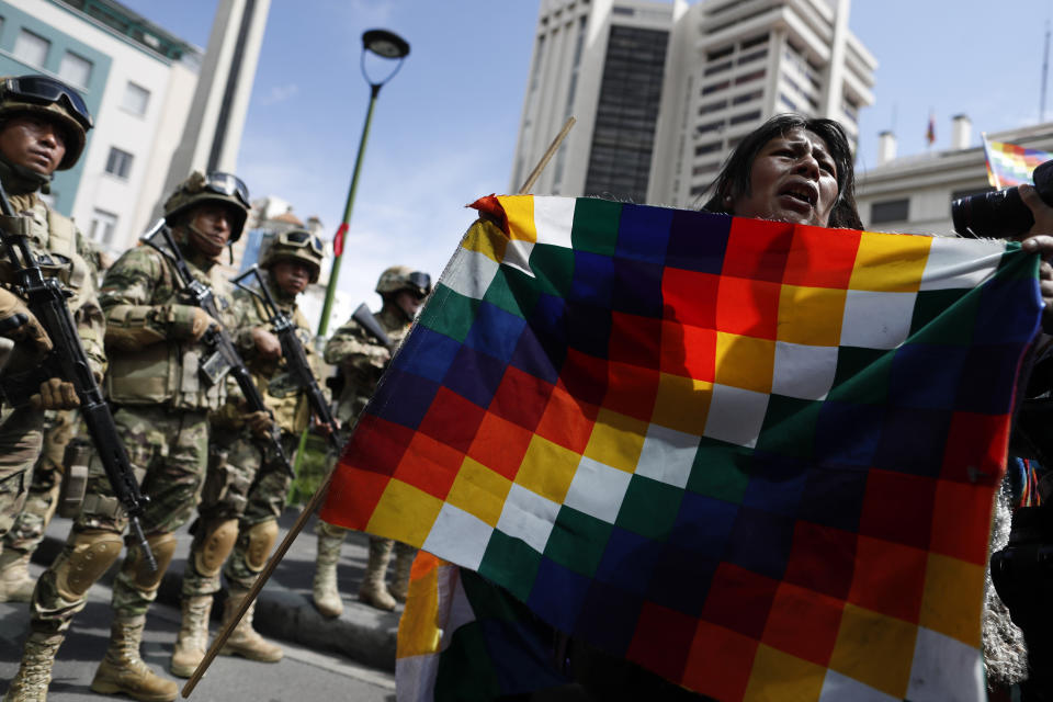 A backer of former President Evo Morales holds a Wiphala flag in front of soldiers blocking a street in downtown La Paz, Bolivia, Friday, Nov. 15, 2019. Bolivia's new interim president Jeanine Anez faces the challenge of stabilizing the nation and organizing national elections within three months at a time of political disputes that pushed Morales to fly off to self-exile in Mexico after 14 years in power. (AP Photo/Natacha Pisarenko)