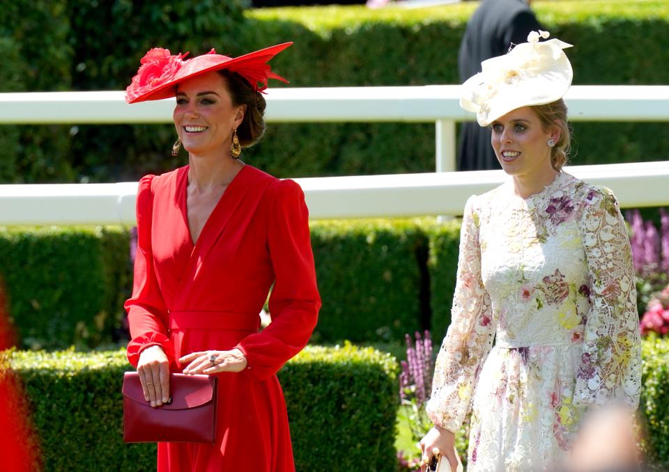 Kate Middleton holding a handbag while wearing a bright red dress and a matching sun hat next to Princess Beatrice, who is wearing a white floral dress and matching sun hat.