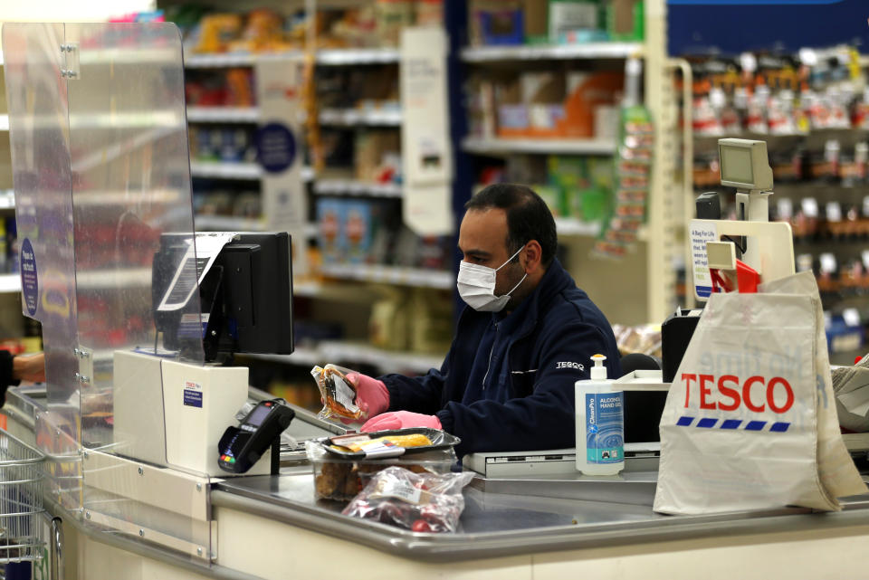 A Tesco supermarket cashier wearing protective face mask and gloves assists a shopper behind a plastic screen in a supermarket in north London as the coronavirus pandemic continues to grow in the UK