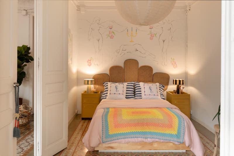 Neutral bedroom featuring matching tables and lamps, paper lantern pendant light, bed with colorful quilt and cane headboard.