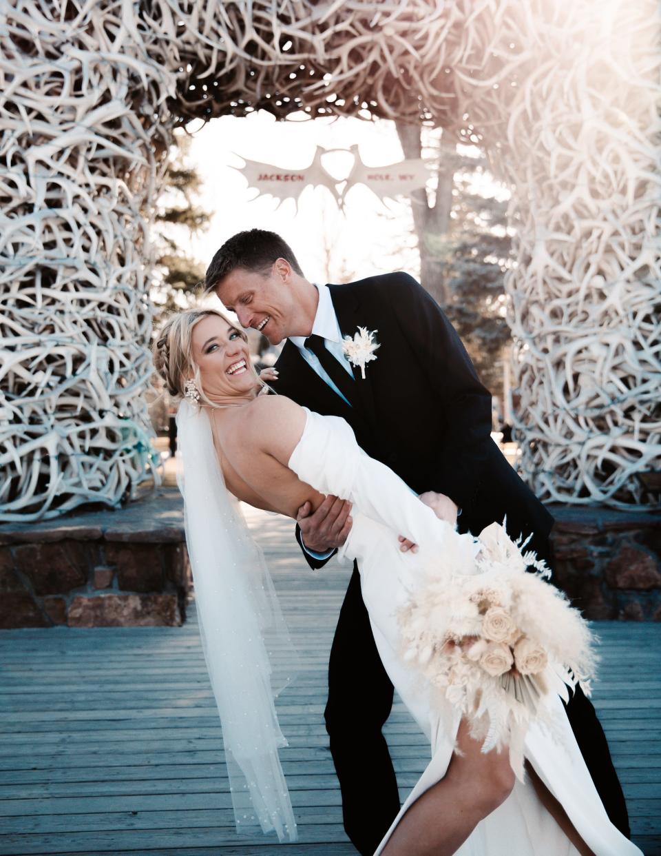 A bride and groom smile in front of an archway as he dips her for a photo.