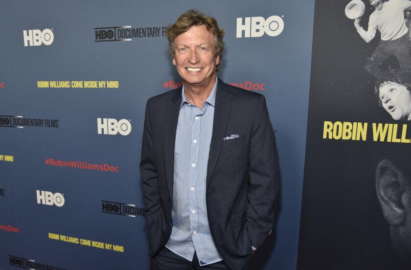 Nigel Lythgoe smiles and poses while wearing a dark blue suit jacket and a light blue dress shirt