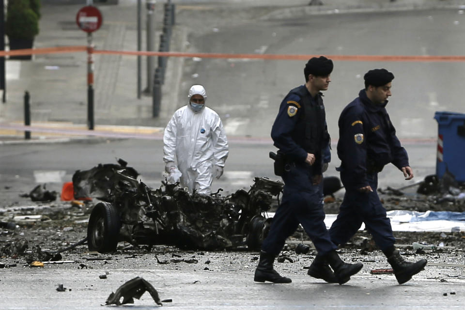 A police bomb disposal expert searches for evidence behind the remains of a car as two policemen walk after a car bomb explosion in central Athens on Thursday, April 10, 2014. A bomb exploded outside a Bank of Greece building in central Athens before dawn Thursday, causing some damage but no injuries. The blast came hours before Greece was to return to the international bond markets for the first time in four years, and a day before German Chancellor Angela Merkel was to visit Athens. (AP Photo/Petros Giannakouris)