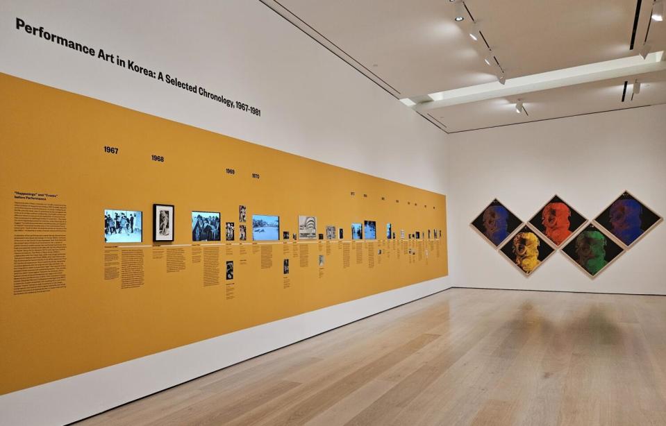 A timeline of still and video images is displayed on a yellow museum wall. Five silkscreen images hang beside it.