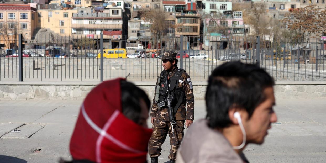 Two people in Kabul pass close to the camera. In the background is a Taliban soldier and a Kabul cityscape.