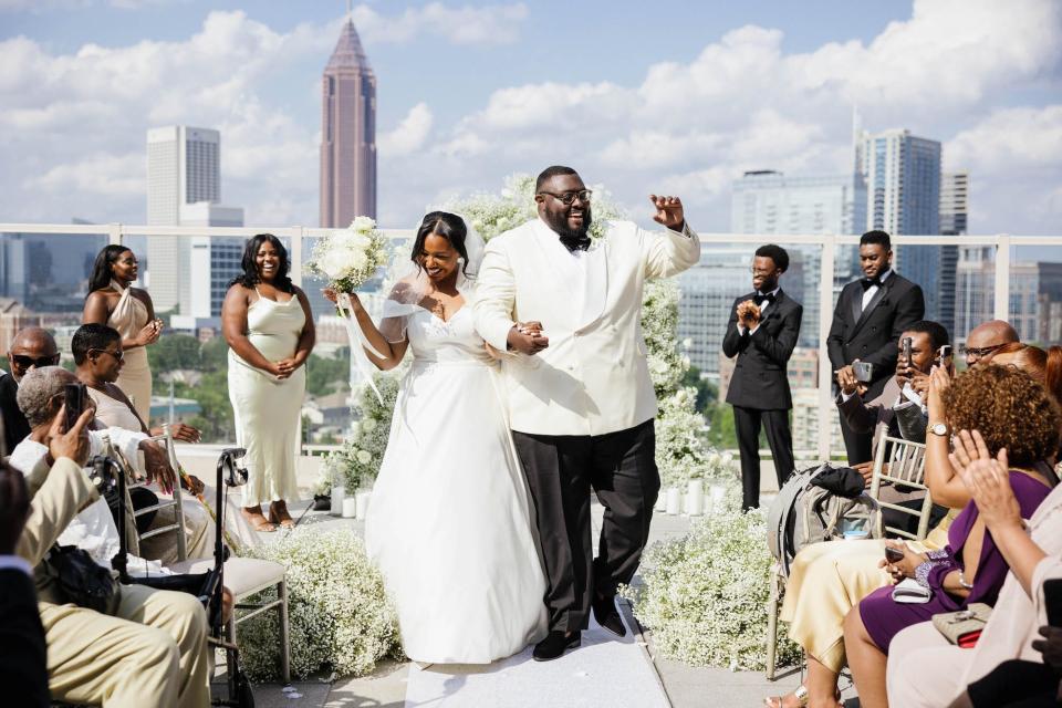 A bride and groom raise their hands in celebration as they leave their wedding ceremony overlooking a city skyline.