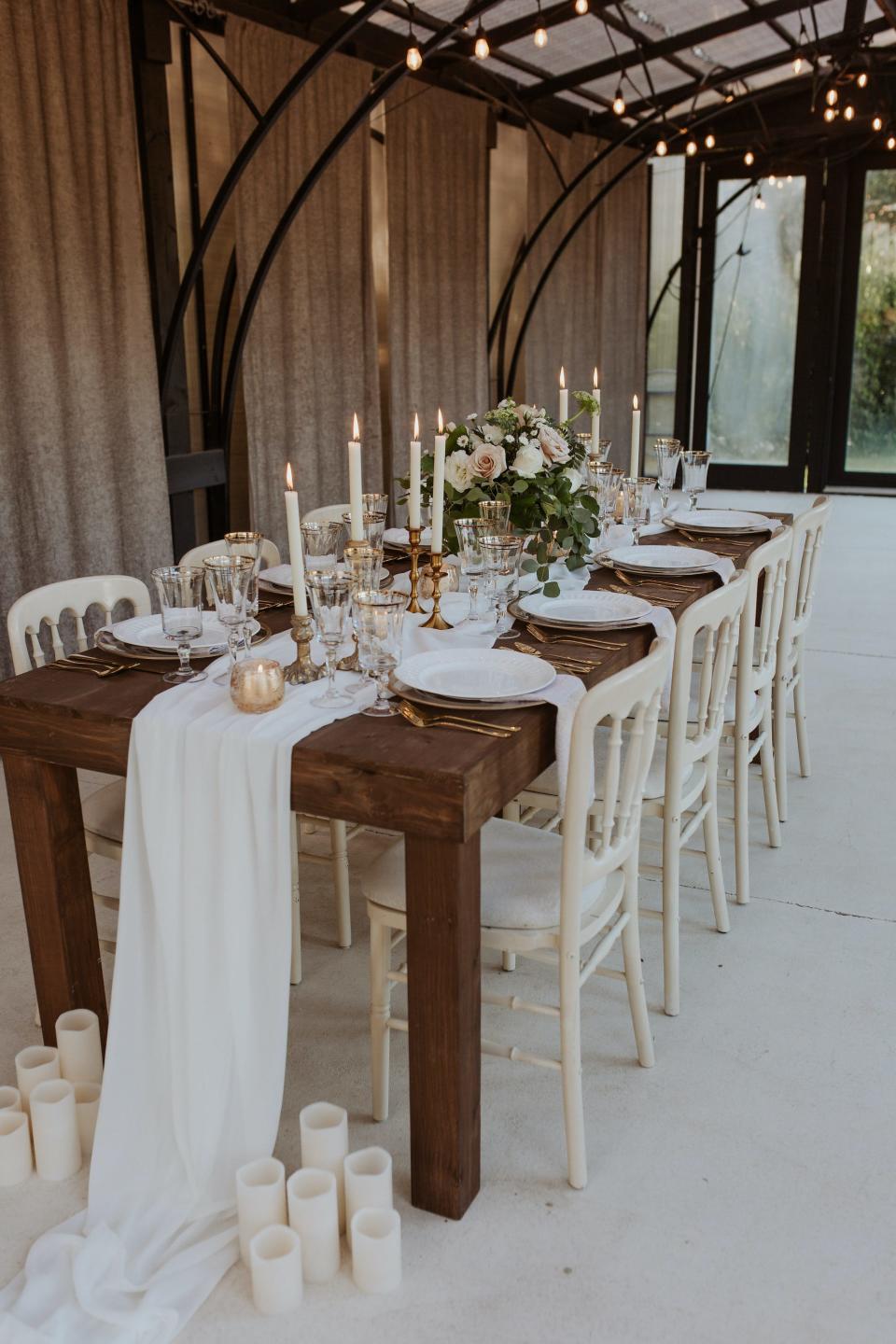 A table with a white runner, candles, and white and gold plates in a room covered in windows.