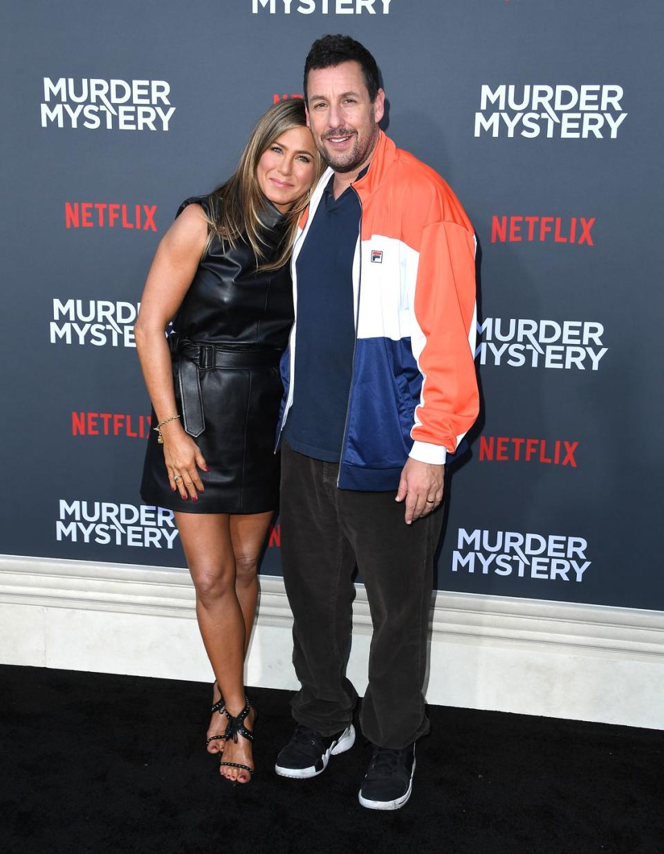 jennifer aniston and adam sandler pose for photos, smiling and standing in front of a black murder mystery movie background