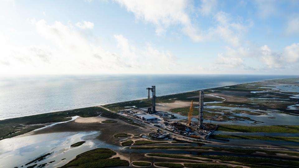 Fernansicht eines Startturms, der eine silberne Rakete über dem Boden hält, mit dem Meer im Hintergrund und Feuchtgebieten rund um die Startrampe