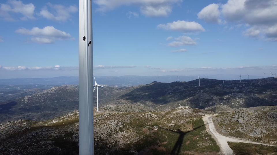 Wide shot of two wind turbine technicians inspecting the blade of a turbine with mountain landscape in the background.