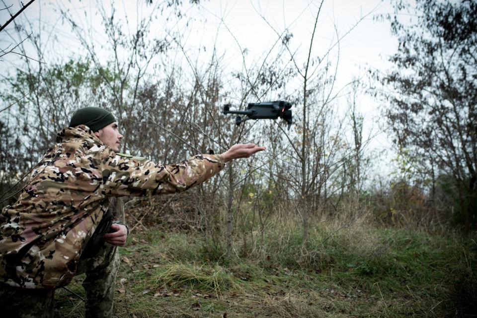 A man wearing camouflage clothing and a green hat extends his hand as a small drone flies away.