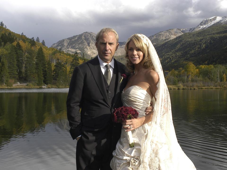 Kevin Costner standing next to Christine Baumgartner at their wedding while standing in front of a large lake with tall trees and mountains in the background.