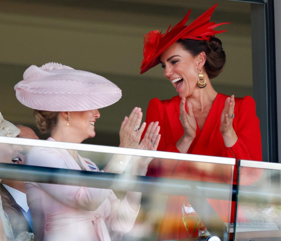 Sophie, the Duchess of Edinburgh, and Kate Middleton smiling and clapping their hands while looking out across a glass balcony.