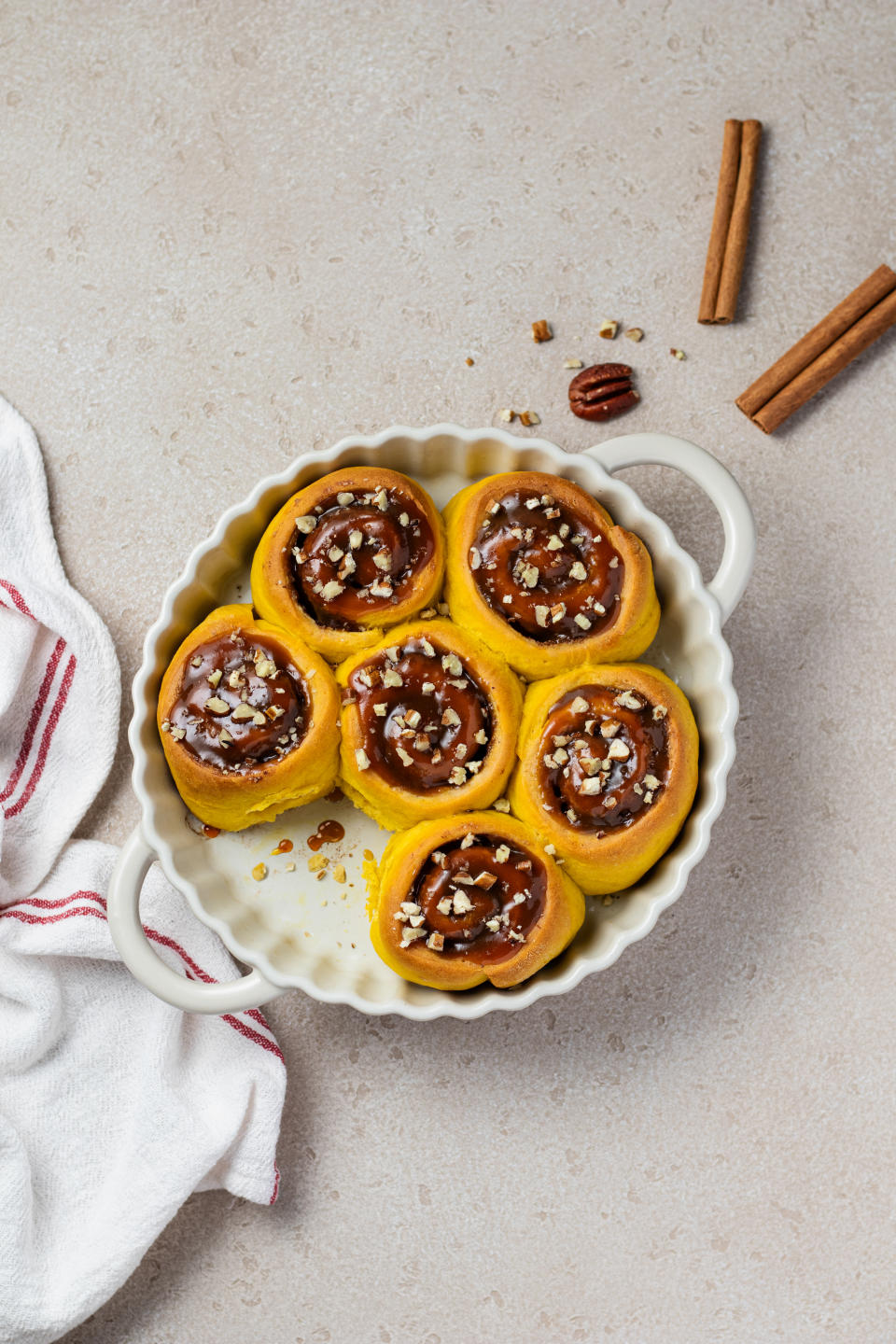 Six cinnamon rolls topped with chopped pecans in a round white baking dish, with two cinnamon sticks and a white cloth beside it