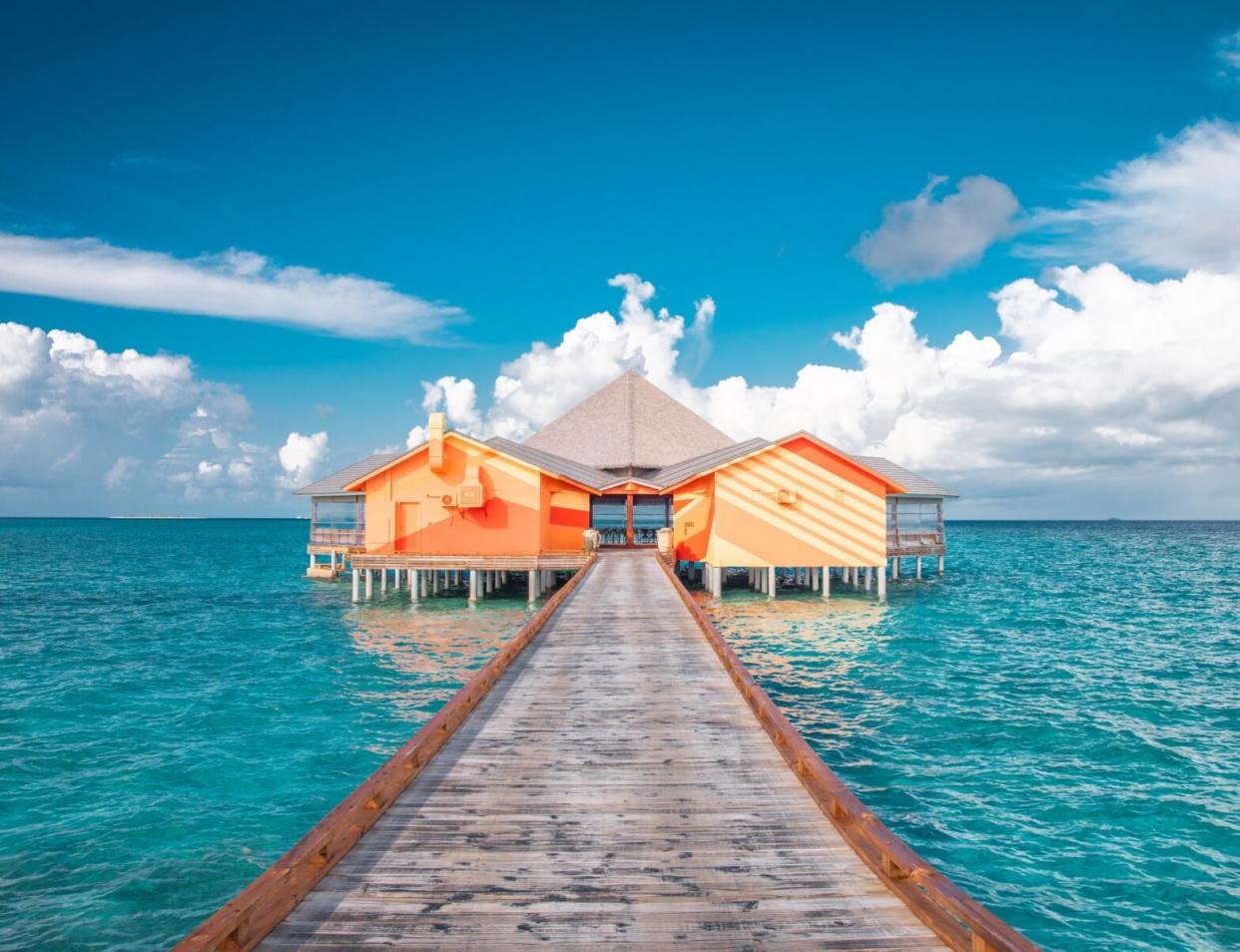 wooden dock leading out to a bright orange twin standing huts over crystal blue waters