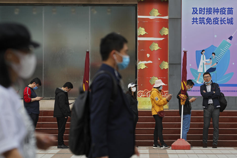 People wearing face masks to help curb the spread of the coronavirus walk by masked residents lining up for COVID-19 vaccine at a vaccination site with a board displaying the slogan, "Timely vaccination to build the Great Wall of Immunity together" in Beijing, Wednesday, April 21, 2021. (AP Photo/Andy Wong)