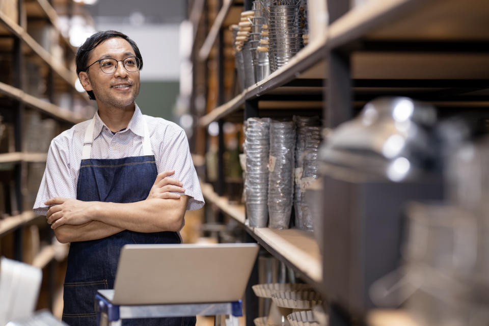 A man in glasses and an apron stands with his arms crossed in a warehouse hallway, smiling and looking to the side, with metal products and a laptop in the foreground