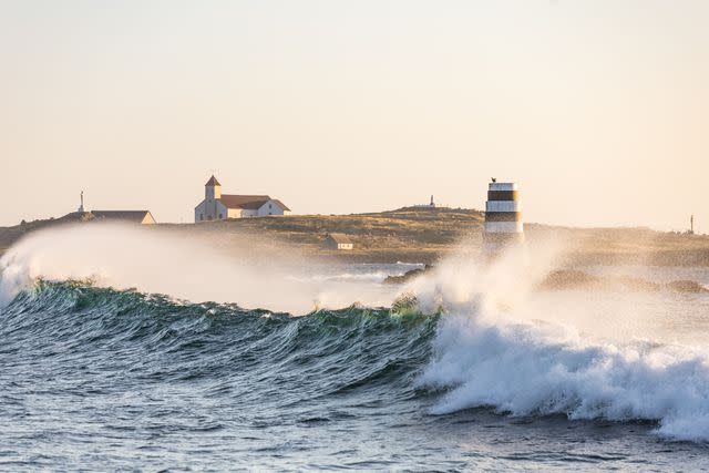 <p>Mathieu Dupuis/Legendary Coasts CVB</p> A wave crashes along the coast of Miquelon near a lighthouse.