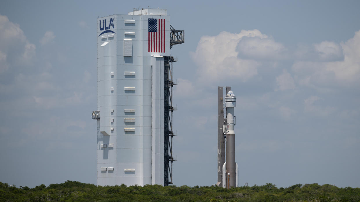 A white and brown rocket stands vertically next to a tall white building. 