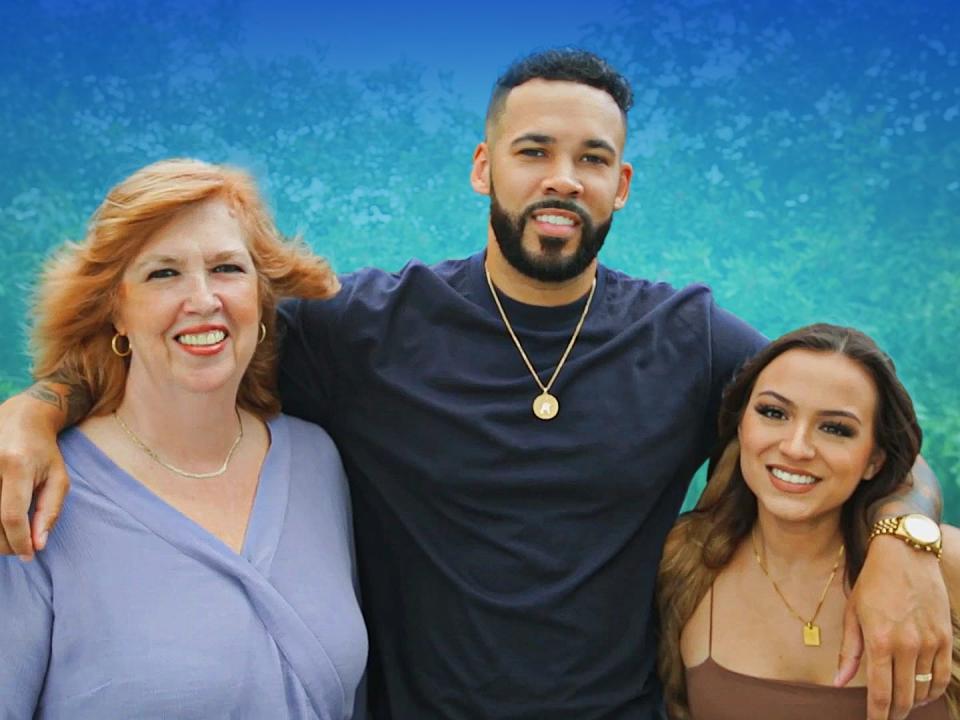 A mother stands next to her son, who has his arms around her and his wife.