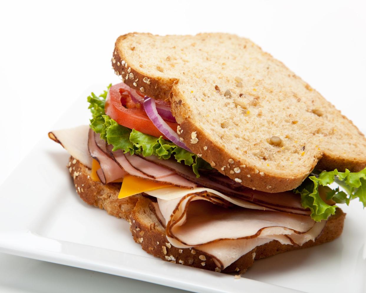 Deli sandwich with ham, cheddar cheese, lettuce, tomato, and onions on a square white porcelain plate on a white table and background