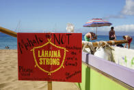 A man sets up an umbrella in front of a Lahaina Strong informational sign, Wednesday, Dec. 6, 2023, Kaanapali Beach in Lahaina, Hawaii. Residents and survivors still dealing with the aftermath of the August wildfires in Lahaina have mixed feelings as tourists begin to return to the west side of Maui, staying in hotels still housing some displaced residents. A group of survivors is camping on the resort beach to protest and raise awareness for better long-term housing options for those displaced. (AP Photo/Lindsey Wasson)