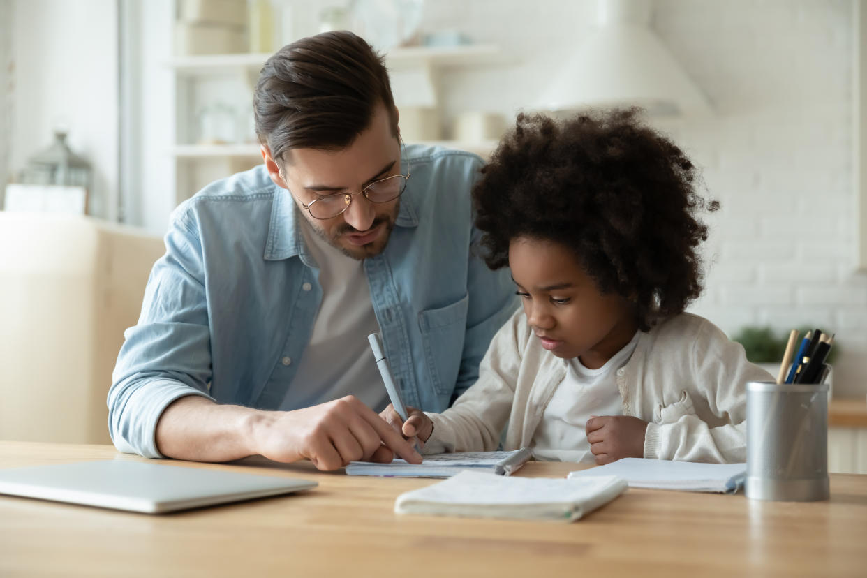 Caring Caucasian father help biracial little daughter with homework at home, loving European dad and small African American girl child study together in kitchen on quarantine, homeschooling concept