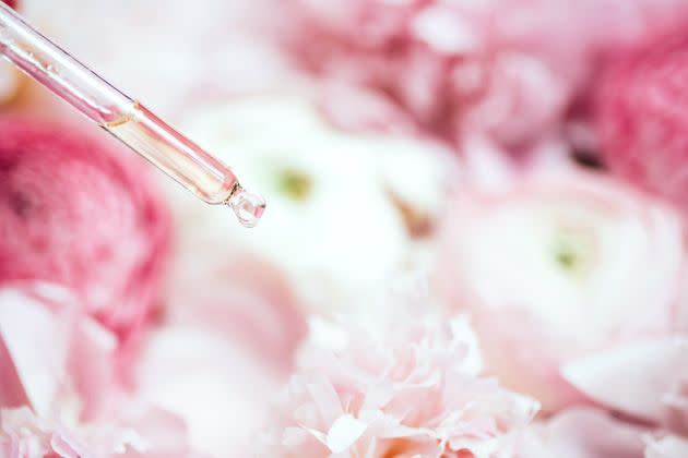 Face serum is dripping from a glass pipette on pink peonies and a ranunculus background. Concept of beauty procedures and body care. Macro photography