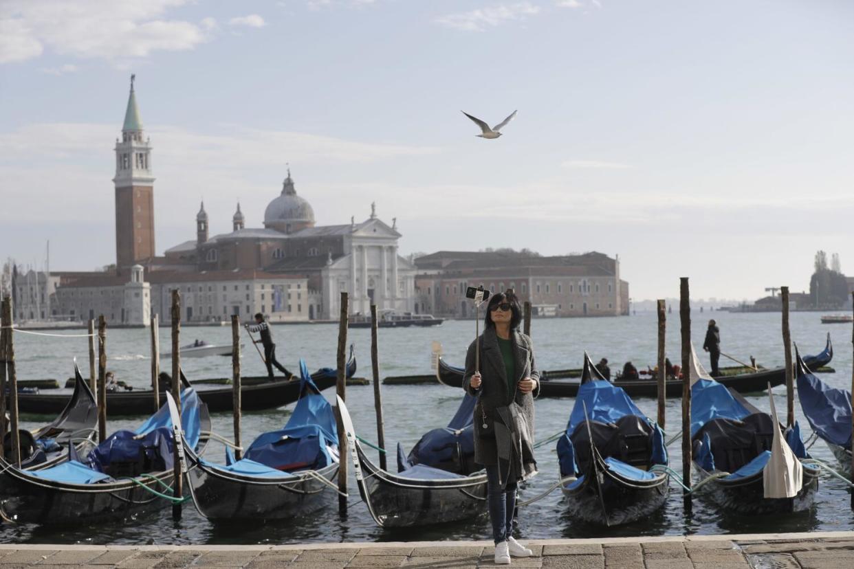 A tourist taking a selfie on a dock in front of several floating gondolas, ornate buildings visible across a stretch of water