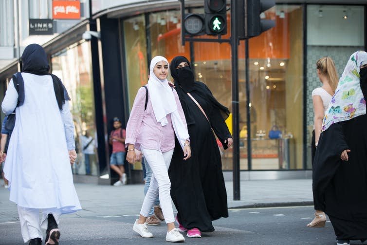 A mother in a black niqab and a daughter wearing white jeans and a white hijab walk down a shopping street.