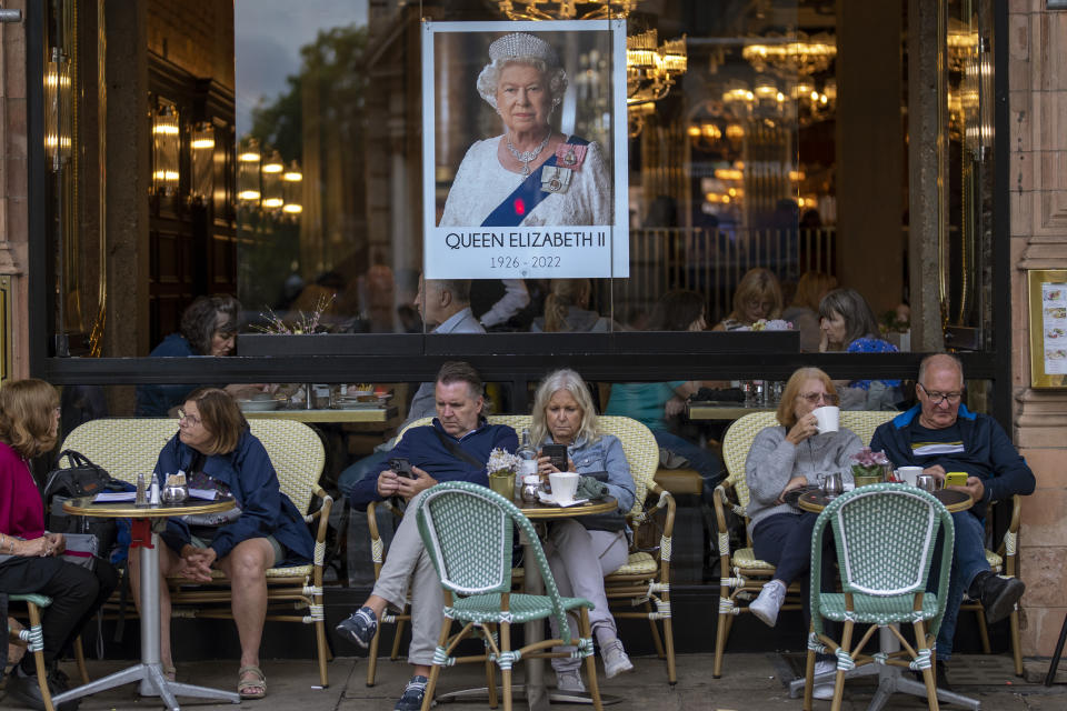 FILE - People sit at a terrace bar next to a portrait of Queen Elizabeth II in central London, Tuesday, Sept. 13, 2022. Hotels, restaurants and shops are packed as royal fans pour into the heart of London to experience the flag-lined roads, pomp-filled processions and brave a mileslong line for the once-in-a-lifetime chance to bid adieu to Queen Elizabeth II. (AP Photo/Emilio Morenatti, File)