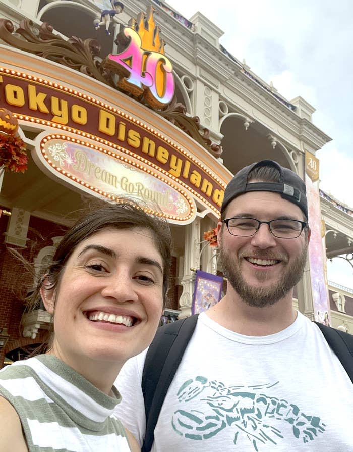 Two people smiling outside Tokyo Disneyland, celebrating its 40th anniversary. The park entrance reads "Tokyo Disneyland" and "Dream Go Round."