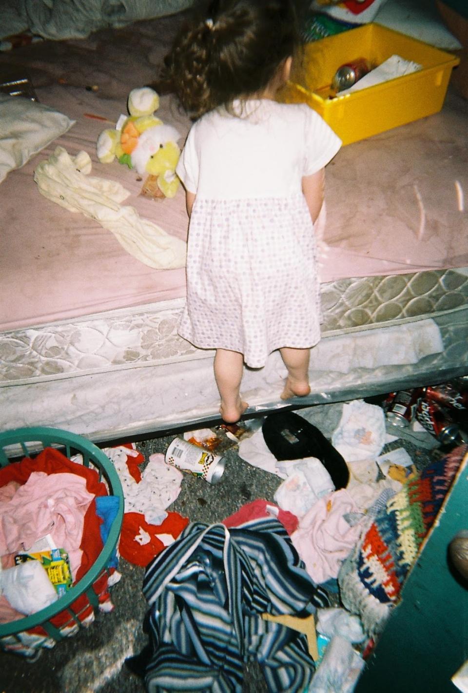 One of Brynlee's sisters standing on the edge of a dirty bed. The floor is covered in clothes and rubbish.