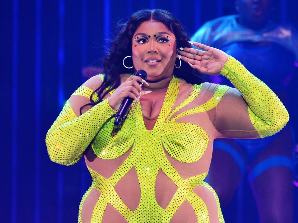 Lizzo speaking into a microphone while raising her hand to her ear and wearing a yellow figure-hugging dress.