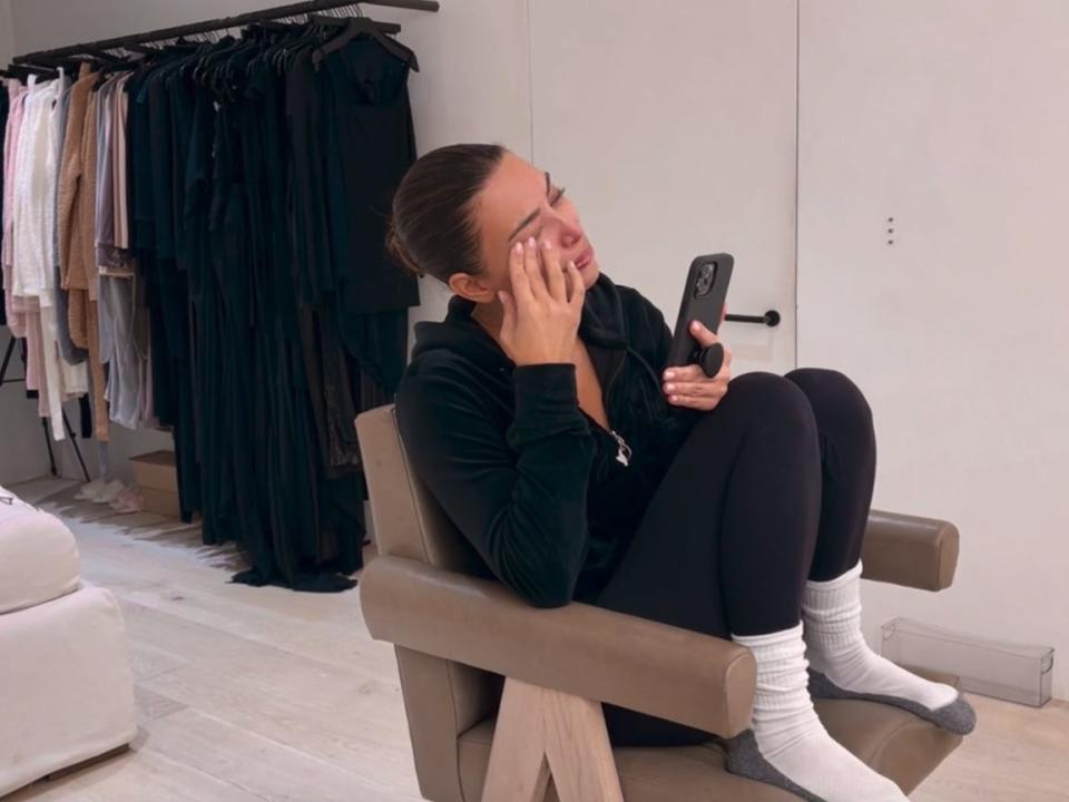 kim kardashian sits in a chair in a wardrobe room, her feet pulled up to the cusion. she's on the phone and is wiping a tear from her eye