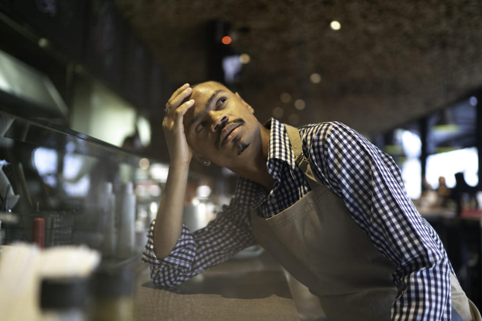 Man in apron leaning on counter, appearing thoughtful, in a café setting