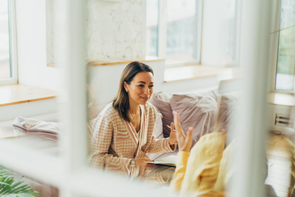 Woman in a casual sweater smiling and gesturing, sitting indoors with a notebook