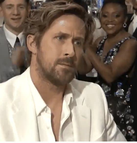 Ryan Gosling, in a white suit, stares ahead with a serious expression while people in the background smile and applaud