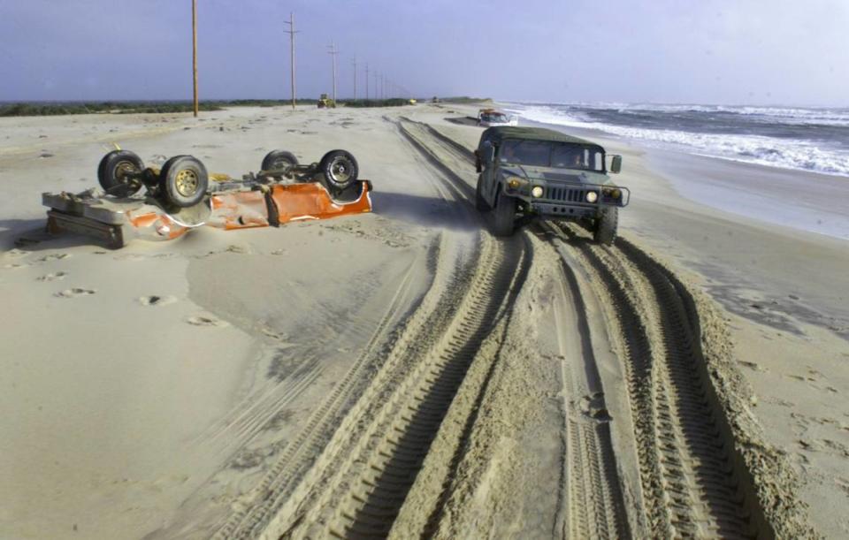 A North Carolina Army National Guard humvee makes its way past an overturned truck partially buried in the sand on a section of what used to be Highway 12 just north of Buxton in 1999.  Heavy surf from Hurricane Dennis overwashed several sections of the road, destroying the dune line and depositing several feet of sand. 