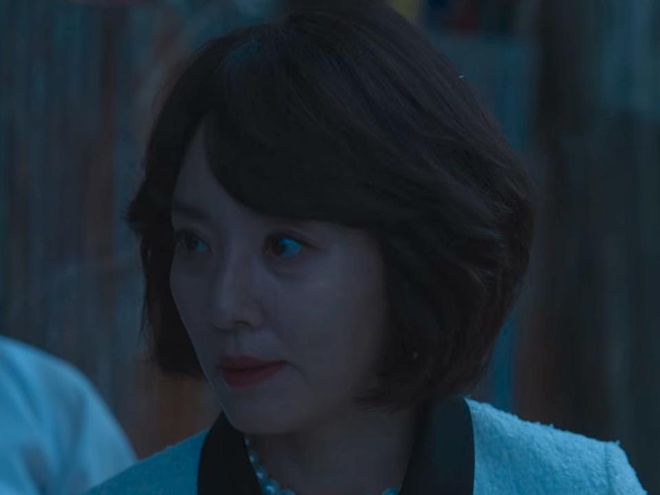 an older middle aged woman with a wavy bob haircut looking to the side at her daughter off-screen. she's wearing a suit, and there's a younger woman visible behind her wearing korean hanbok