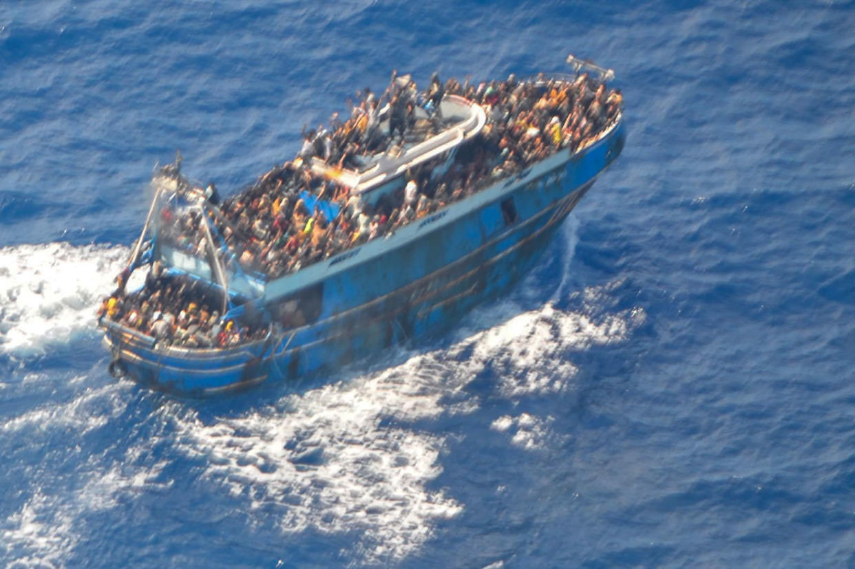 A grainy photograph that shows a large blue battered fishing boat in the ocean filled with people