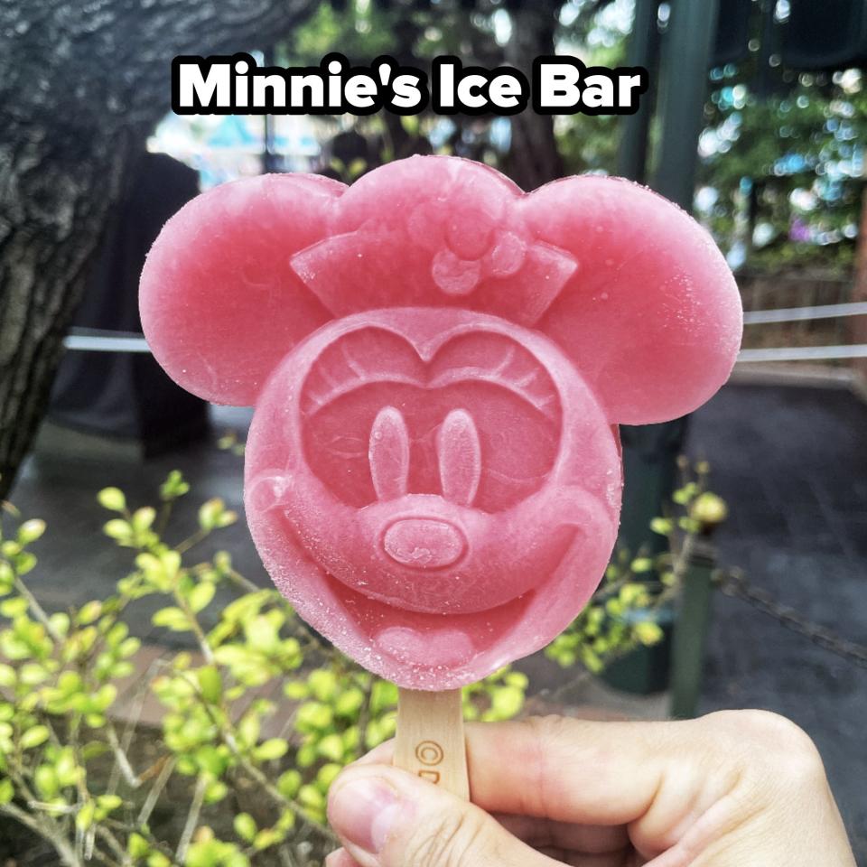 A person holds a Minnie Mouse-shaped ice cream bar on a stick in what appears to be a park