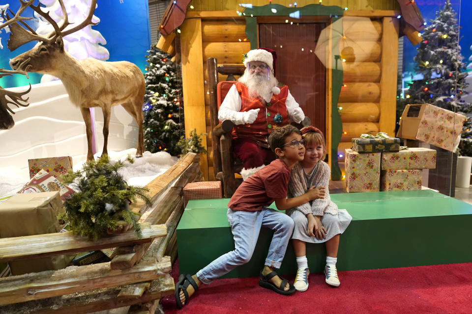 Theo and Sophy Morris, visiting from Hawaii with their family, pose for a photograph with Santa Claus, who is sitting behind a transparent barrier, at Bass Pro Shops in Miami on Nov. 20, 2020. In this socially distant holiday season, Santa Claus is still coming to towns (and shopping malls) across America but with a few 2020 rules in effect. (AP Photo/Lynne Sladky)